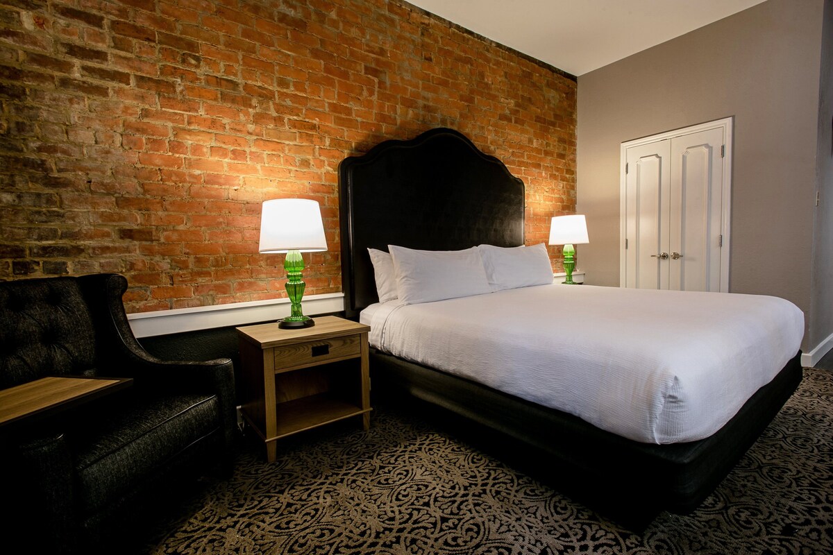 Une chambre d’hôtel avec un mur en briques, un lit recouvert de draps blancs, deux lampes de chevet, une table de chevet en bois, un fauteuil noir recouvert de tissu et une moquette à motifs.