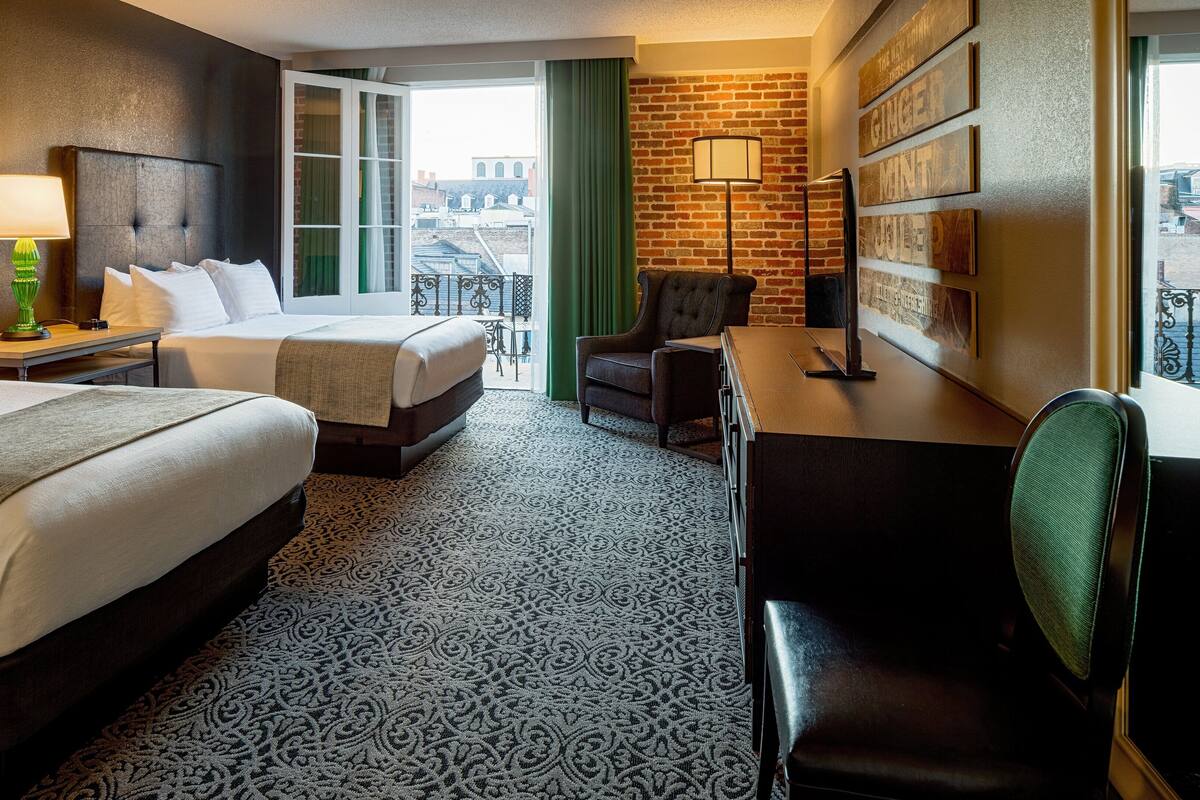 Ein Hotelzimmer mit zwei Betten, einem Schreibtisch, einem Stuhl, einer Lampe und Blick auf die Stadt durch das Fenster.