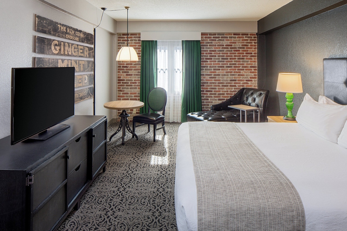 Een hotelkamer met een bed, een televisie, een bureau, een stoel, een lamp en een bakstenen muur.