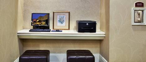 A hotel reception area with a flat-screen TV, a framed picture, a printer, and a 'House Phone' sign. There are two leather ottomans in front of a white counter.