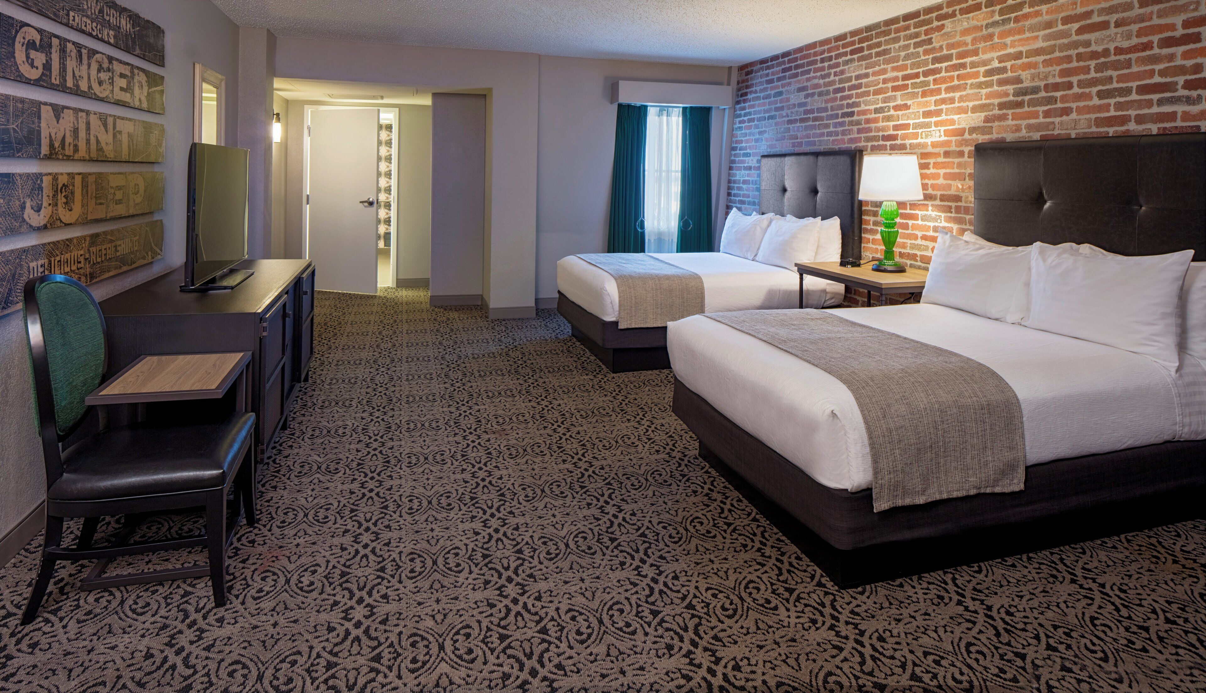 A hotel room with two beds, a desk, and a chair. There is a brick wall and a patterned carpet.