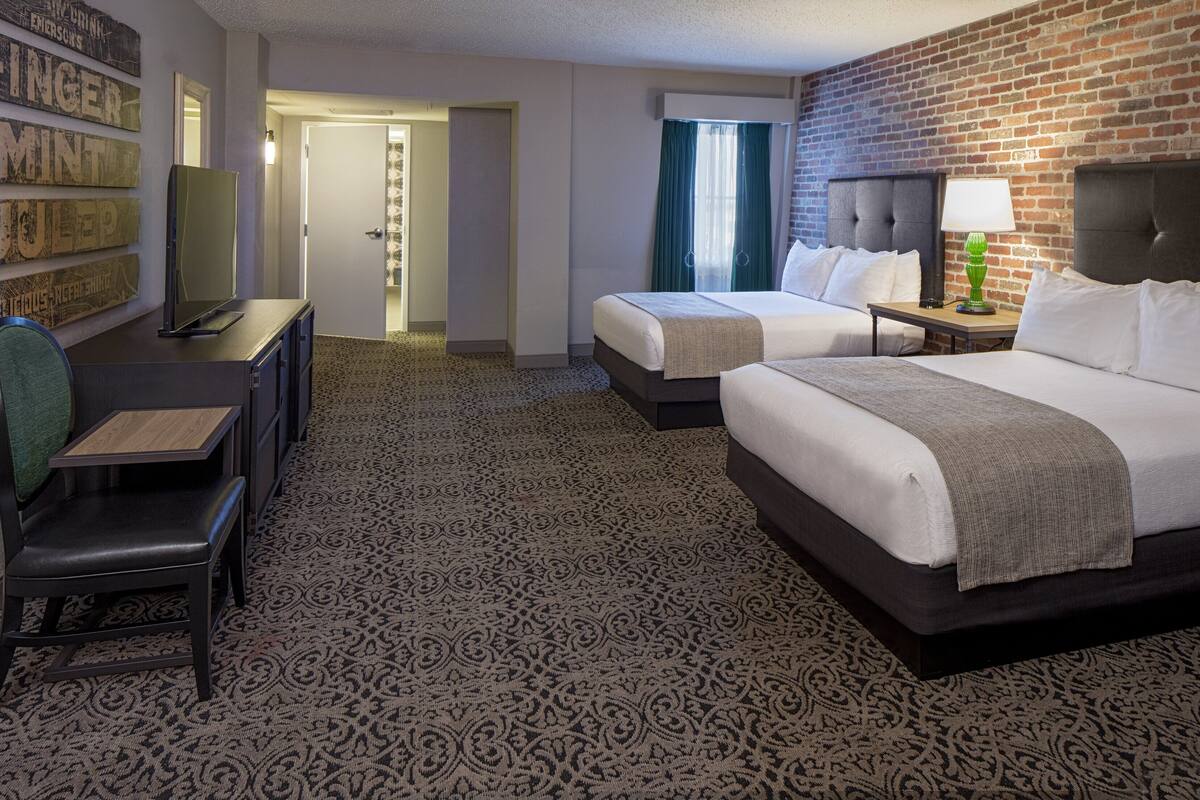 Ein Hotelzimmer mit zwei Betten, einem Schreibtisch und einem Stuhl. Eine Backsteinwand und ein gemustertes Teppich sind ebenfalls vorhanden.