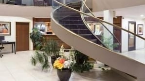 Un hall d’hôtel moderne avec un escalier en verre incurvé, un comptoir d’accueil et des plantes d’intérieur.