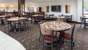 Une salle à manger d'hôtel avec plusieurs tables rondes et des chaises, une cuisine à l'arrière-plan et un grand téléviseur accroché au mur.