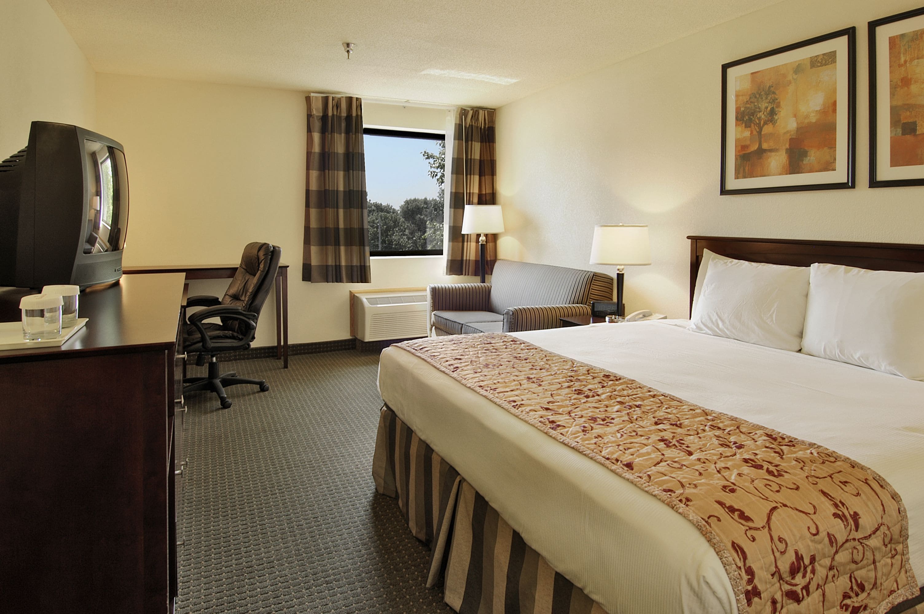 Een hotelkamer met een bed, een bureau met een televisie, een stoel, een raam met gordijnen en twee wandlampen.