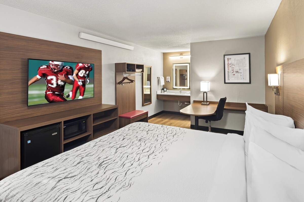 A hotel room with a bed, a desk, a chair, a TV, a microwave, a mini-fridge, a closet, and a bathroom with a sink and mirror.