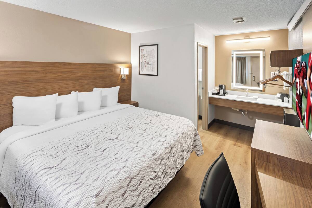 Hotellihuone, jossa on suuri sänky, puinen pääty, yöpöytä lampun kanssa, kylpyhuone peilillä, pesualtaalla ja suihkutilalla.