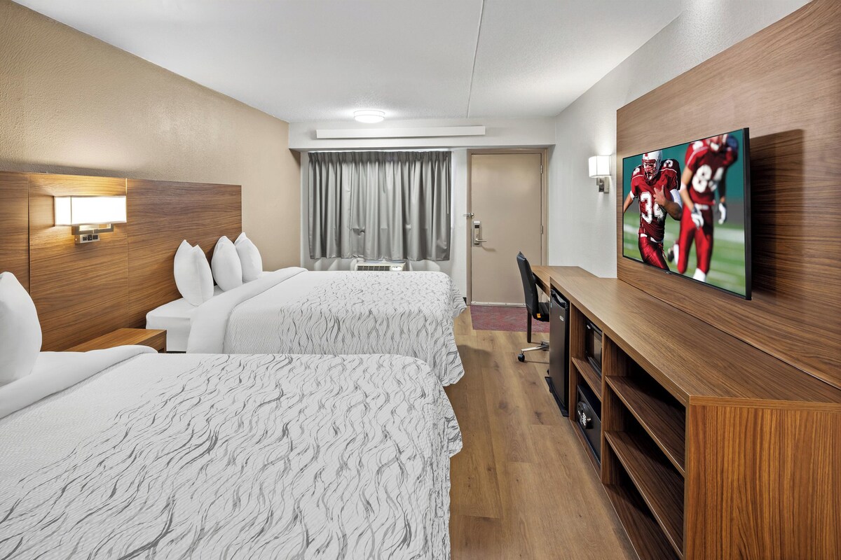 A hotel room with two beds, a TV on the wall, a desk, and a window with curtains.