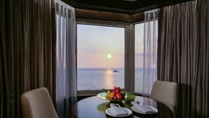 A dining table with a fruit bowl and napkins, facing a window with a view of the sunset over the water. - Diamond Hotel Philippines (Manila)