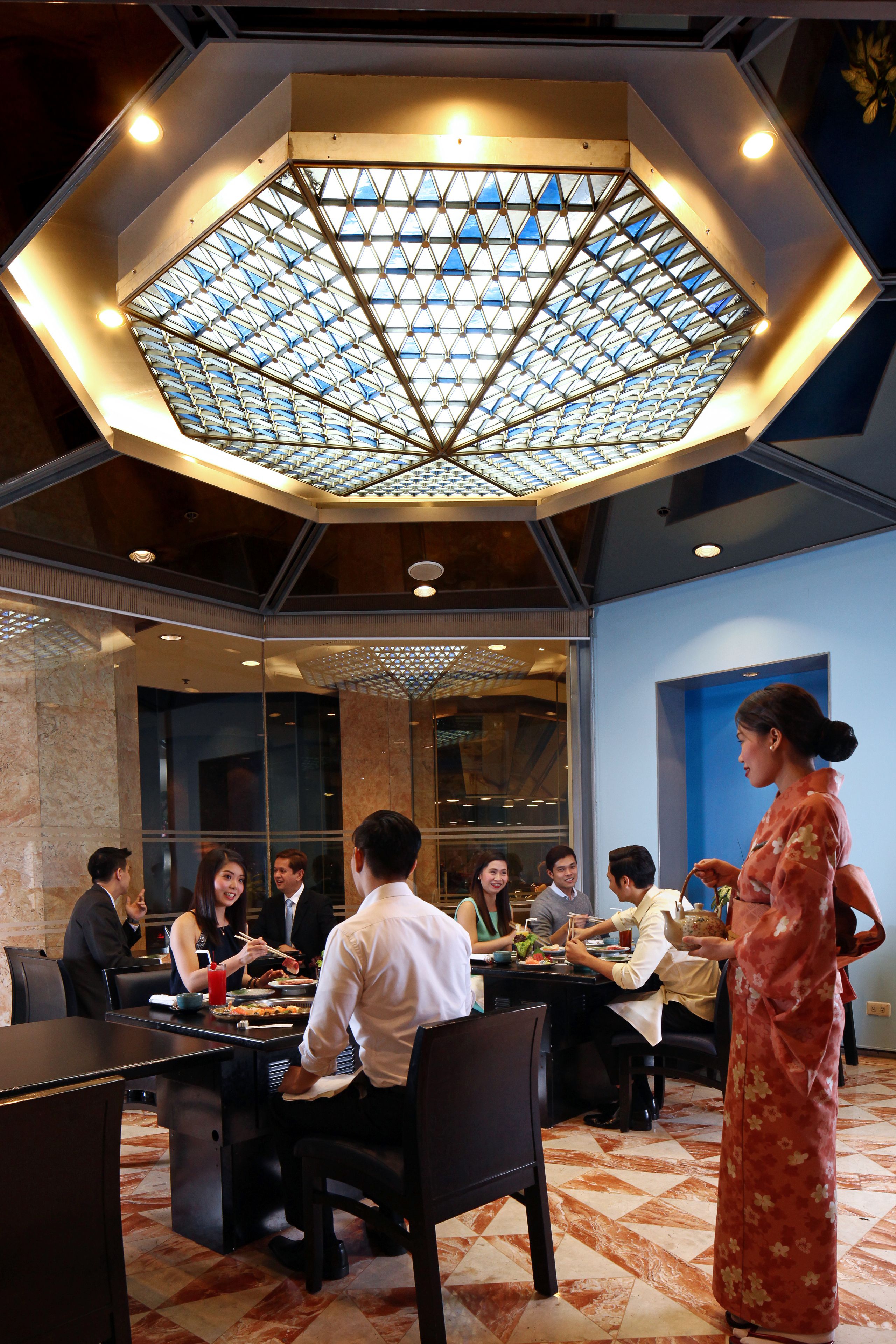 Un comedor con un diseño geométrico en el techo, mesas dispuestas para grupos, y una persona vestida con traje tradicional parada cerca.