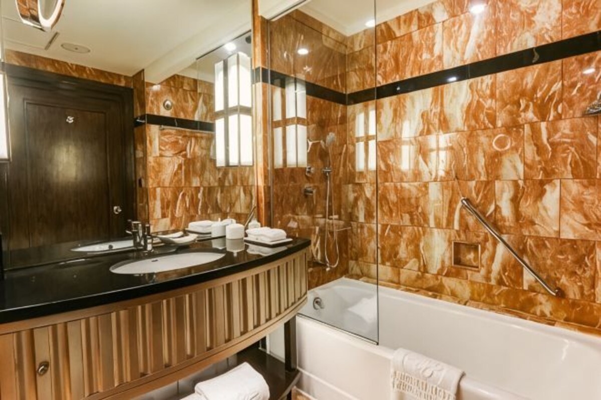 A bathroom with a marble wall, a large mirror, two sinks, a bathtub, and a glass shower enclosure.