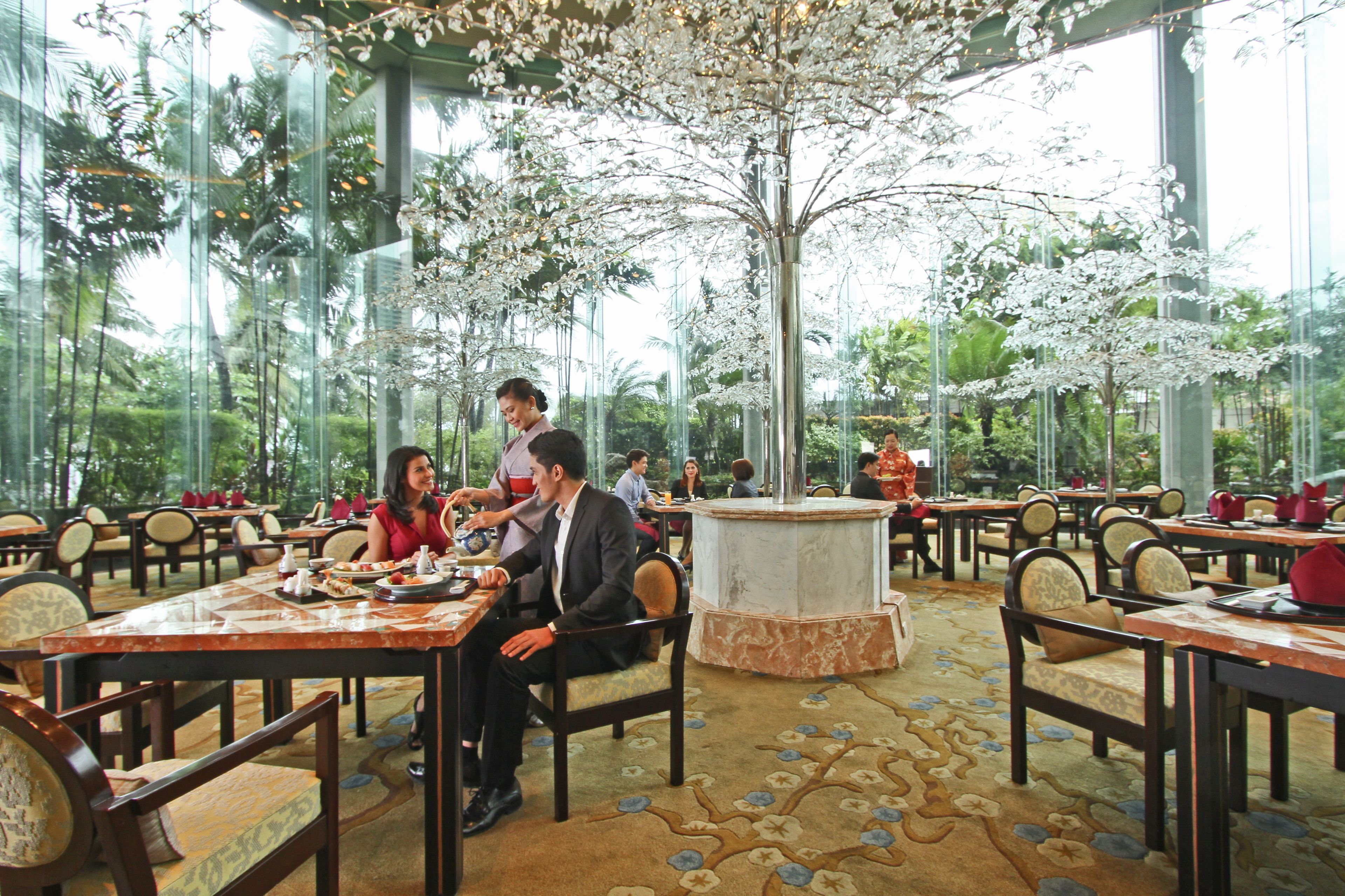 Un restaurante con varias mesas y sillas, ventanas amplias y un árbol decorativo en el centro.