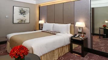 A hotel room with a large bed, two bedside lamps, a bedside phone, a small table with a vase of red flowers, and a large mirror.
