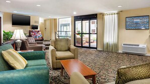 Un hall d’hôtel avec un tapis à motifs, un espace salon avec des canapés et des fauteuils, un téléviseur, un comptoir de réception et une grande fenêtre avec des rideaux.
