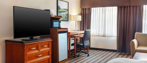 Habitación de hotel con televisor de pantalla plana, escritorio con silla, refrigerador pequeño, cama y ventana con cortinas.