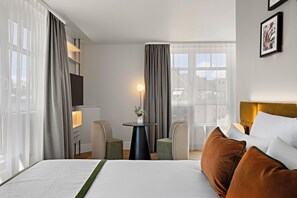 A hotel room with a bed, a small round table, two chairs, a TV, and a window with curtains.