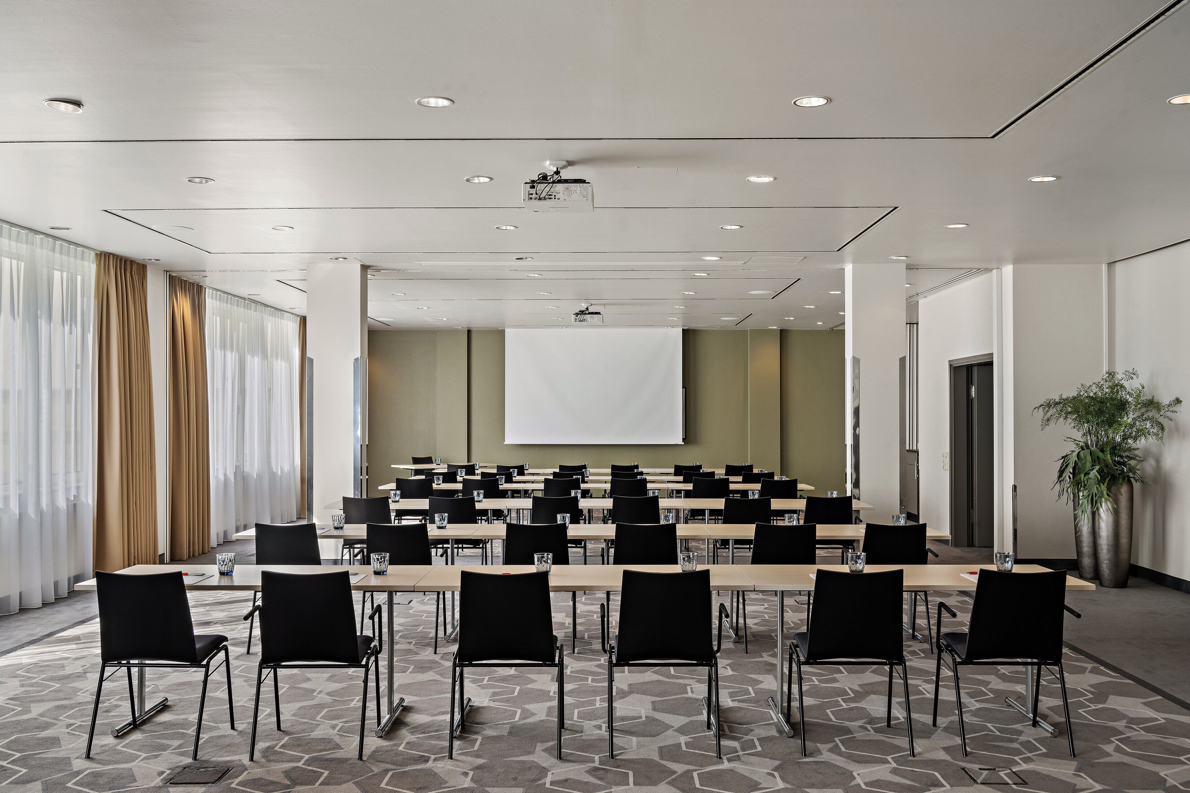 Un salón de conferencias con una larga mesa, sillas negras, una pantalla de proyector y un amplio ventanal con cortinas.