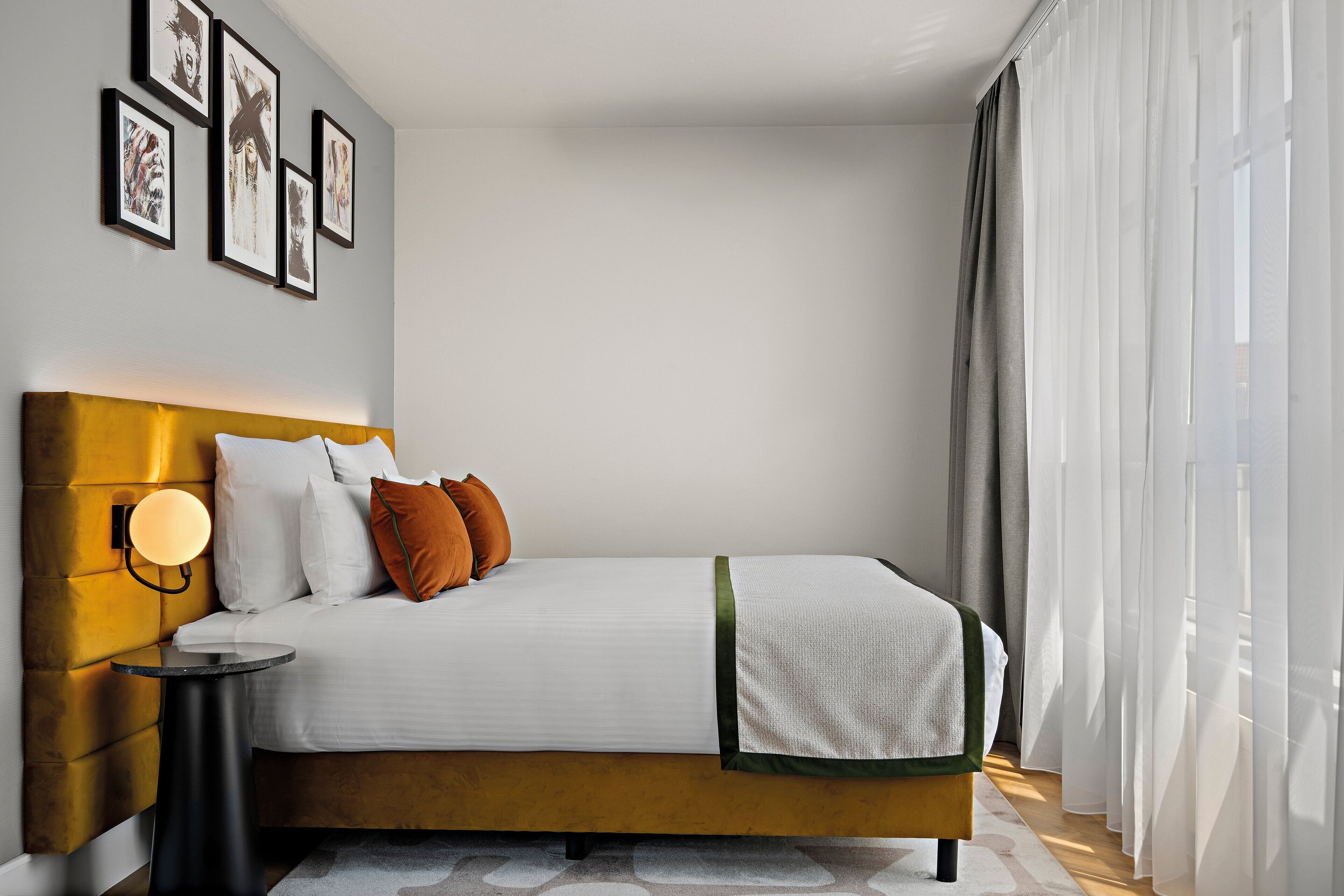 A modern hotel room with a large bed, a bedside table with a lamp, and framed artwork on the wall.