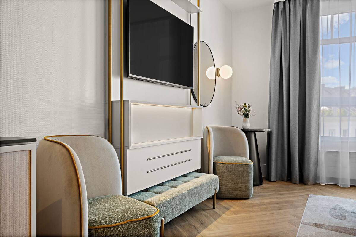 Ein modernes Hotelzimmer mit einem großen Flachbildfernseher, einer Couch, einer Bank, einem kleinen Tisch mit einer Vase und Blick auf die Stadt durch große Fenster.