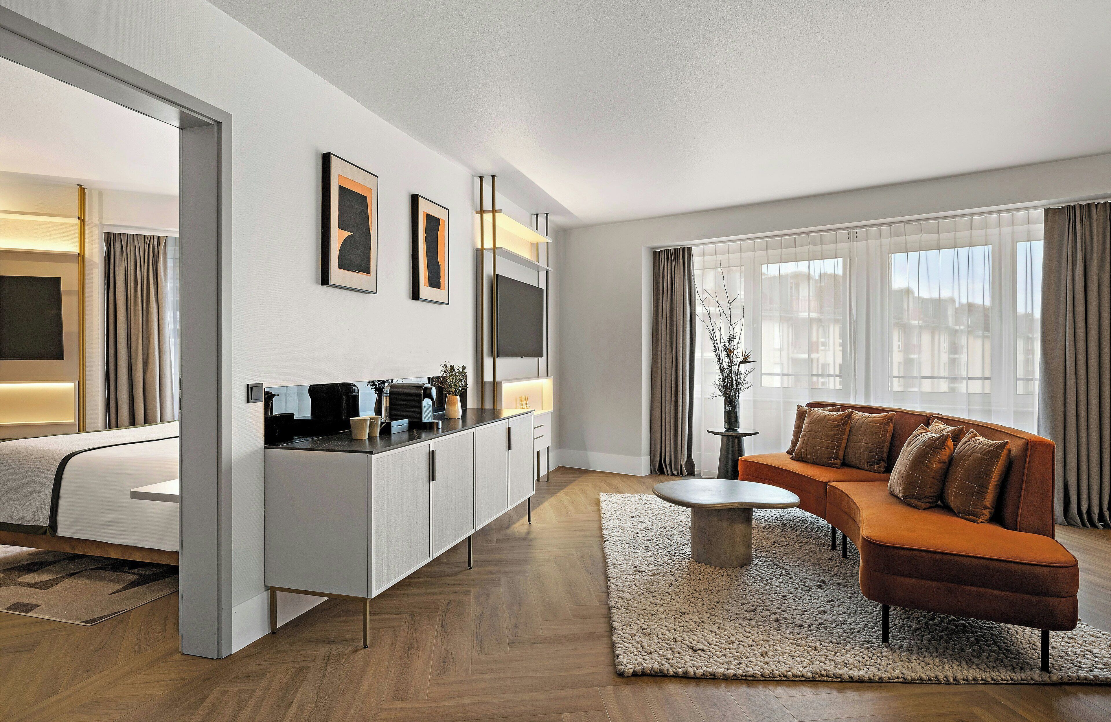 Una habitación de hotel moderna con cama, sofá, mesa de centro, televisor y un ventanal con cortinas.