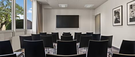 A conference room with black chairs arranged in rows, a large black screen on the wall, and a window with a view of trees and a building.
