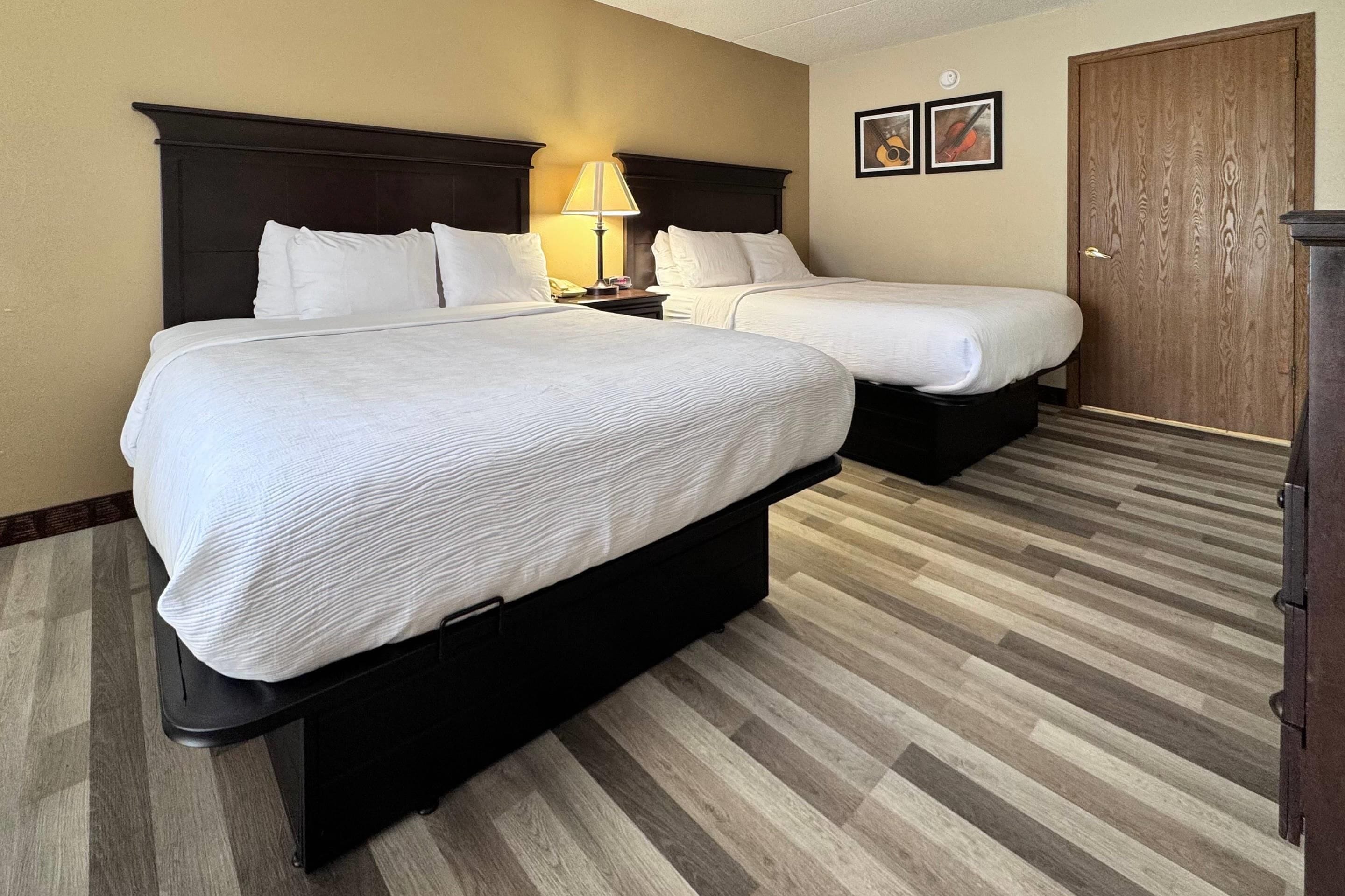 A hotel room with two beds, a nightstand with a lamp, a wooden door, and a framed picture on the wall.