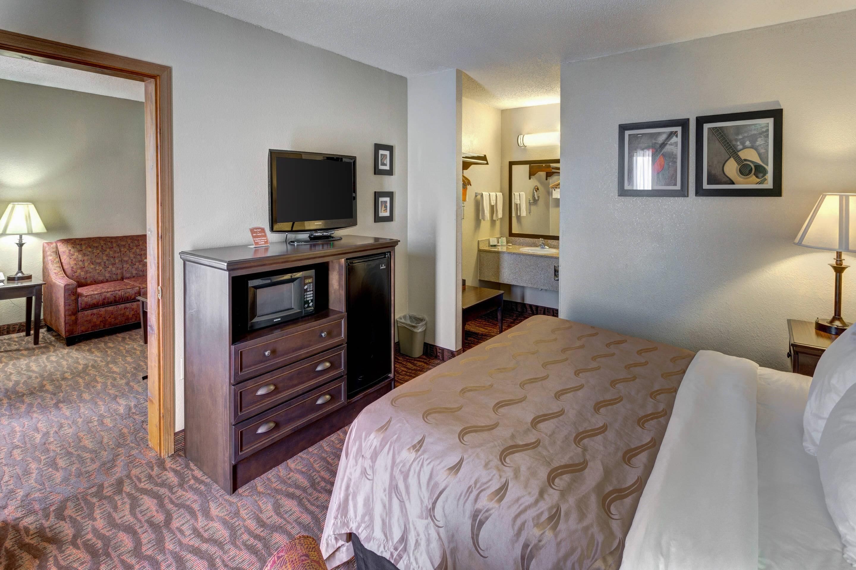 A hotel room with a bed, a television, a desk with a lamp, a chair, and a bathroom with a mirror and towel rack.