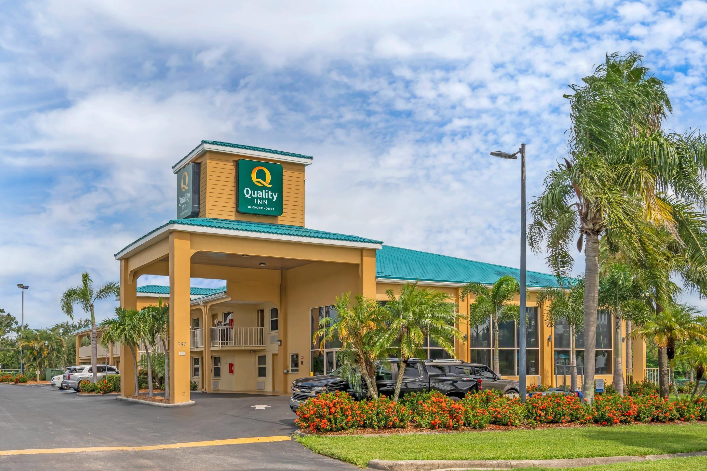 Un hotel Quality Inn con techo verde, un letrero y un estacionamiento con coches y palmeras.