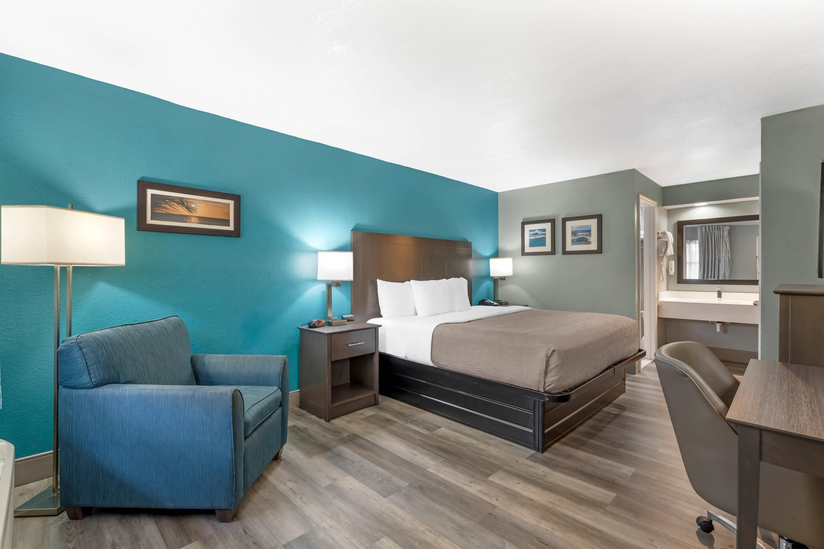 A hotel room with a bed, a desk, a chair, a nightstand, and a lamp.