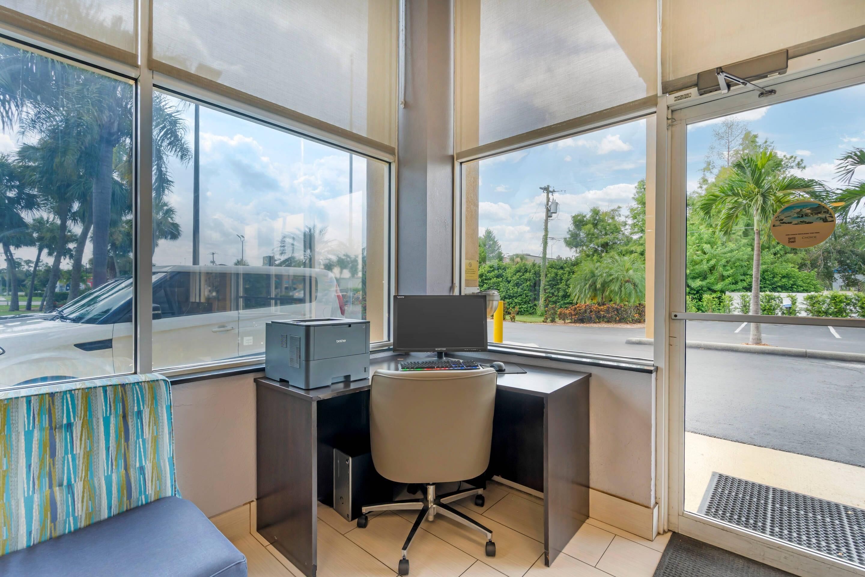 A modern office space with a desk, chair, and large windows offering a view of palm trees and a road.