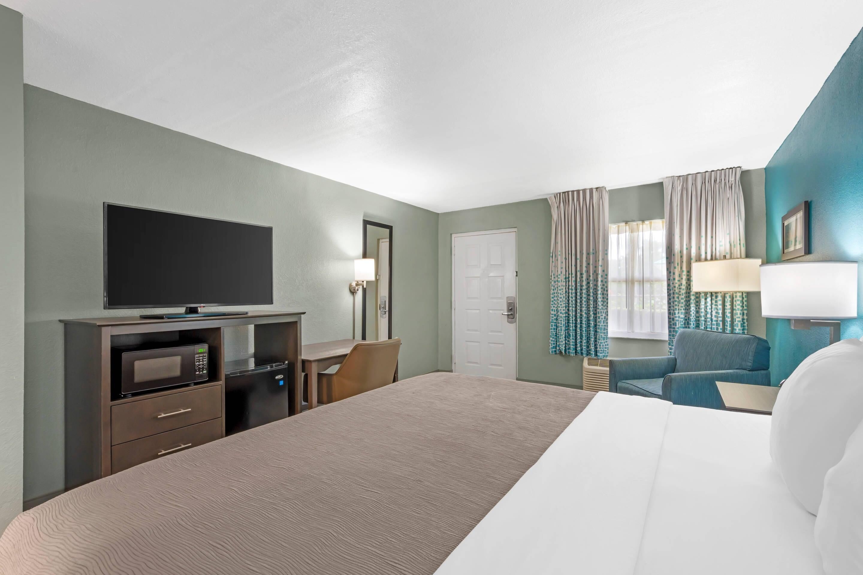 Quarto de hotel com cama, escrivaninha com computador, televisão, micro-ondas e uma cadeira.