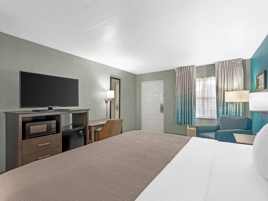 Quarto de hotel com cama, escrivaninha com computador, televisão, micro-ondas e uma cadeira.