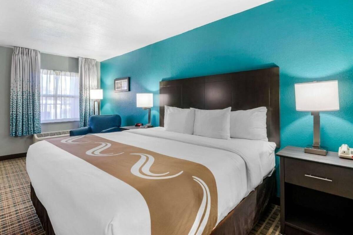 A hotel room with a large bed, a nightstand with a lamp, a chair, and a window with curtains.