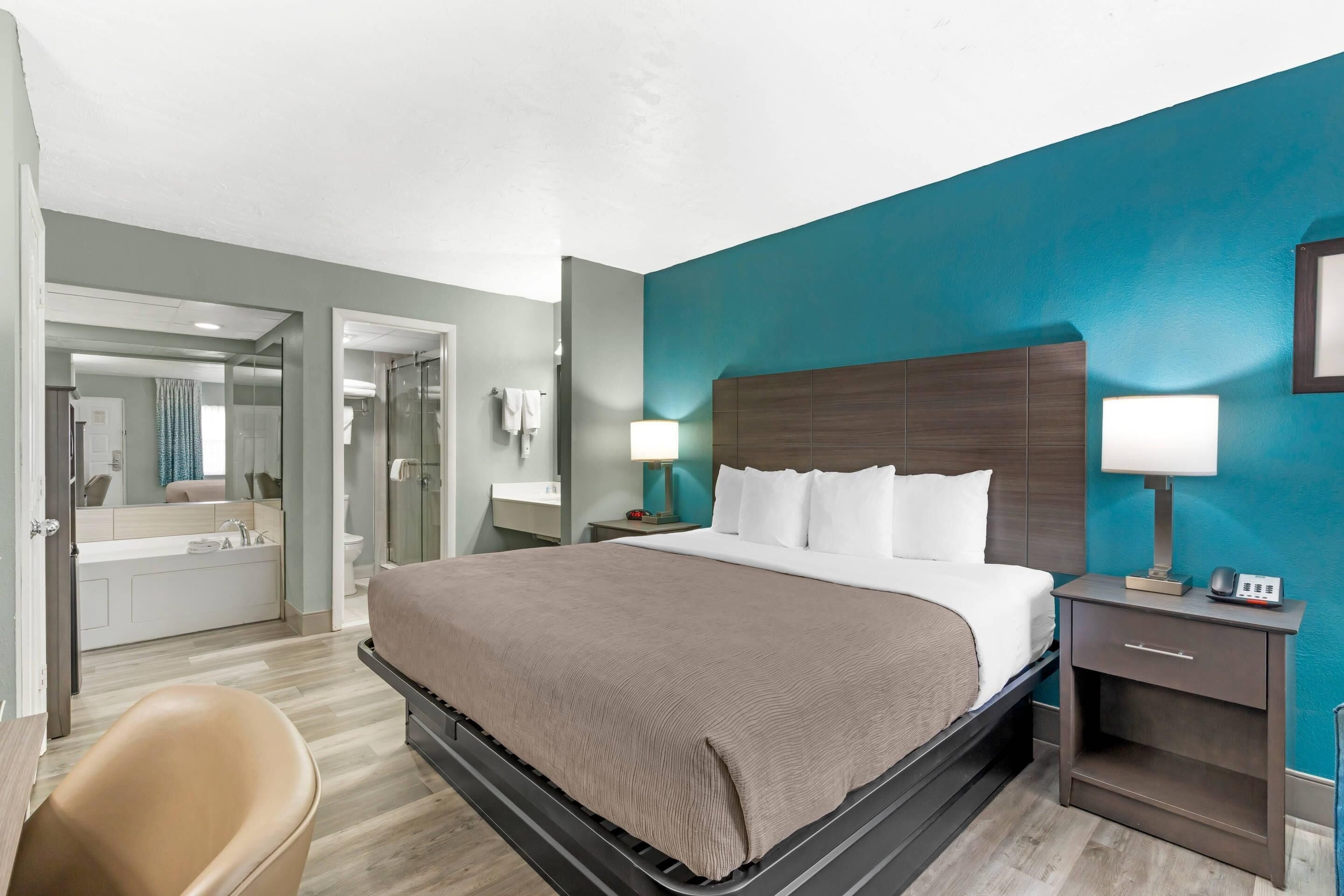 Habitación de hotel con cama, mesita de noche, lámpara y vista al baño.