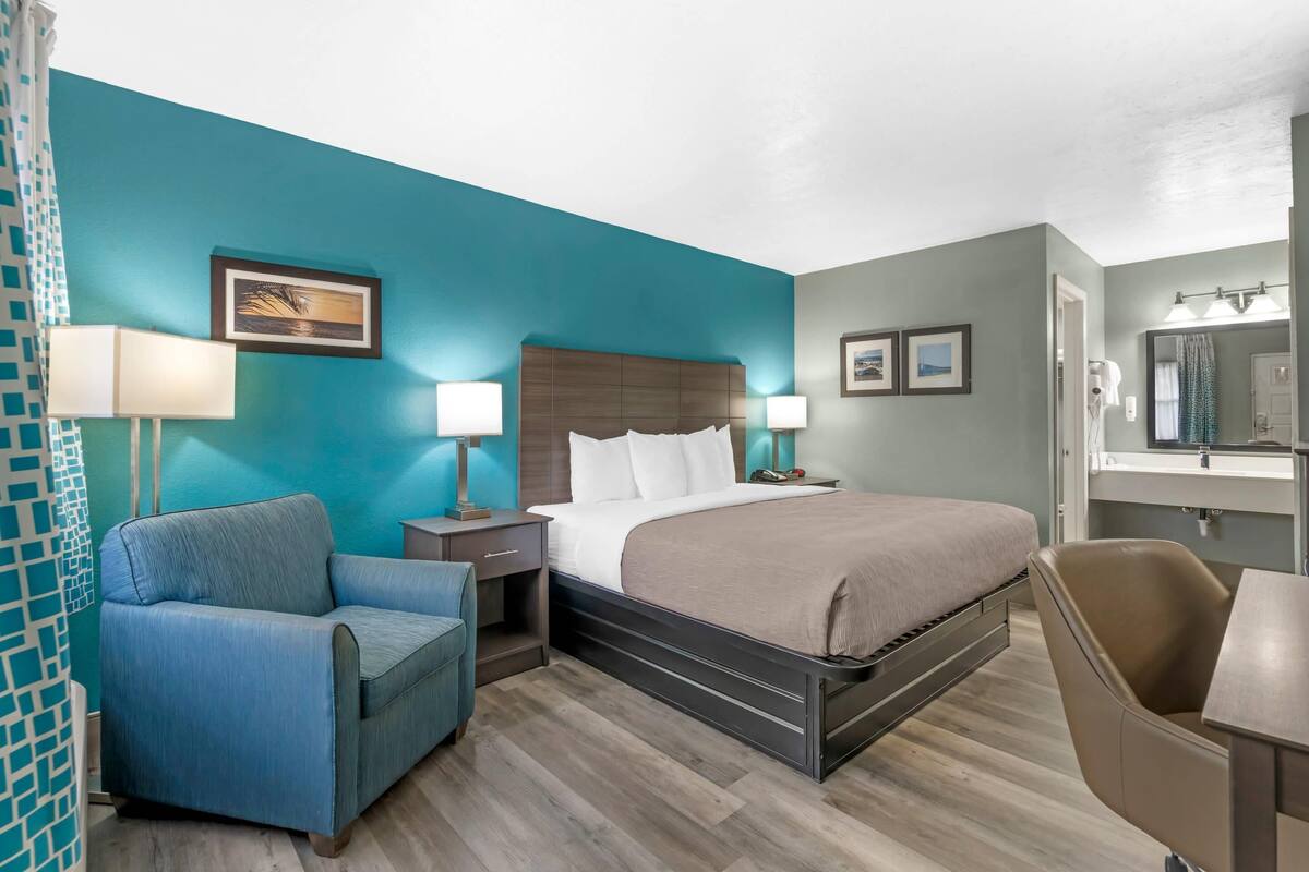 A hotel room with a bed, a desk, a chair, a nightstand, and a bathroom.
