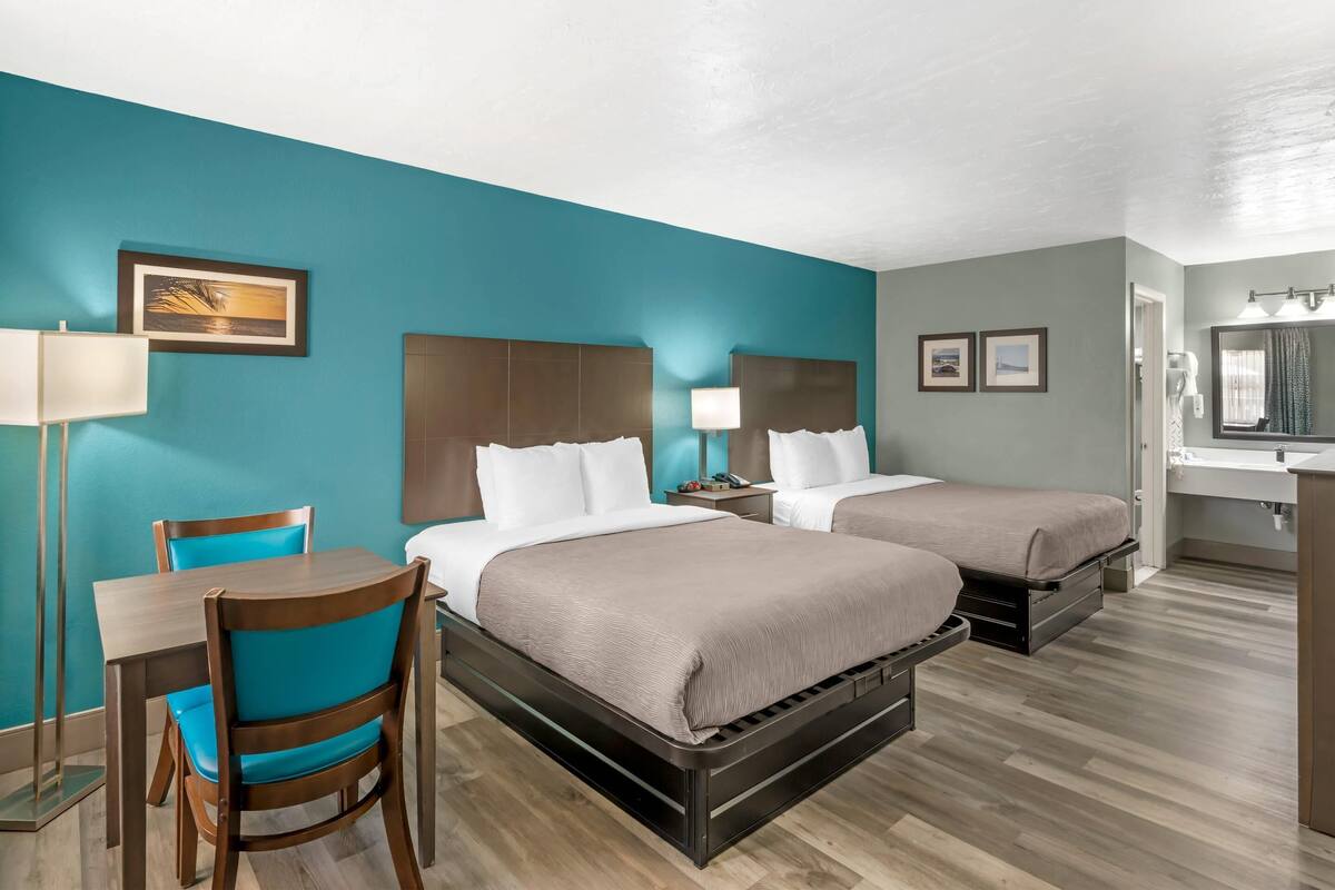 A hotel room with two beds, a desk, and a chair. There is a painting on the wall and a lamp on the desk.