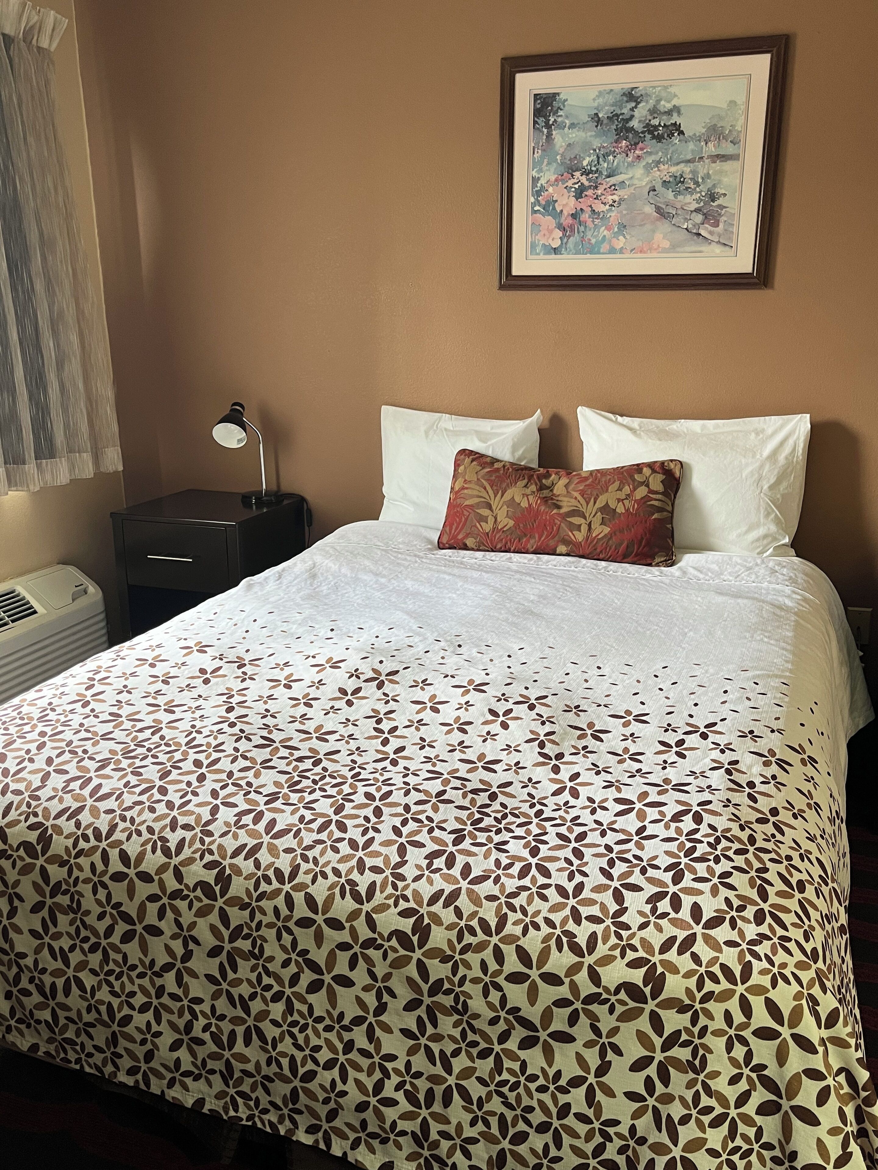 A neatly made bed with a floral-patterned bedspread, a single decorative pillow, and a framed landscape painting on the wall.