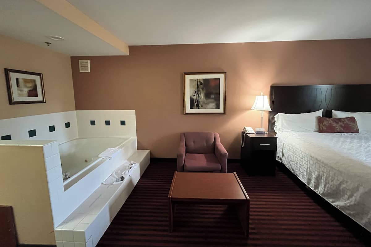 A hotel room with a bed, a chair, a table, a nightstand, a lamp, and a bathtub.
