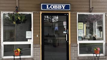 Welcome to the lobby, featuring a black door with a glass panel and two potted plants.