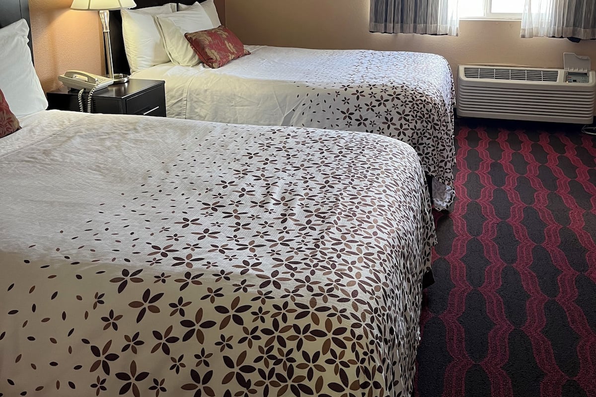 A hotel room with two beds, a bedside table with a lamp, a framed picture on the wall, and a window with curtains.