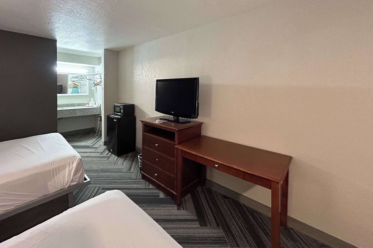Ett hotellrum med två separata sängar, ett skrivbord i trä med en tv, ett litet kylskåp och ett badrum som syns genom en öppen dörr.