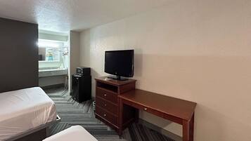 A hotel room with two single beds, a wooden desk with a television, a small refrigerator, and a bathroom visible through an open door.