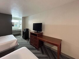 A hotel room with two single beds, a wooden desk with a television, a small refrigerator, and a bathroom visible through an open door.