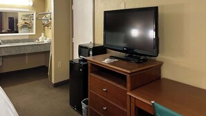 A hotel room with a wooden desk, a flat-screen TV, a microwave, a mini-fridge, a trash bin, a wooden chair, a mirror, a sink, and a towel rack. - Motel 6 Ada, OK (Ada)