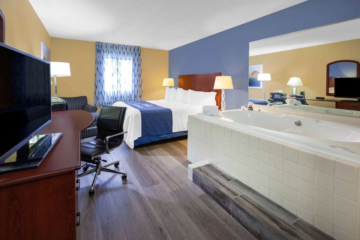 A hotel room with a bed, a desk with a computer, a chair, a large bathtub, and a television.