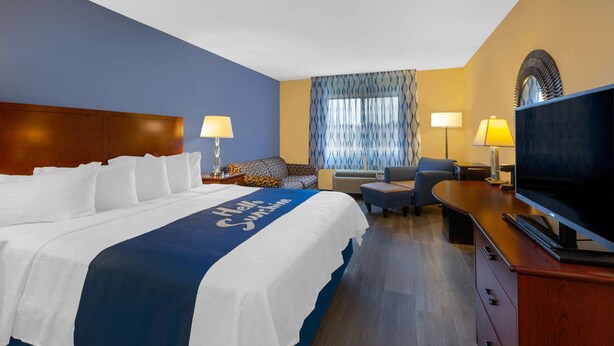 A hotel room with a large bed, a desk with a TV, a sofa, and a window with curtains. - Days Inn by Wyndham Milan Sandusky South (Milan)