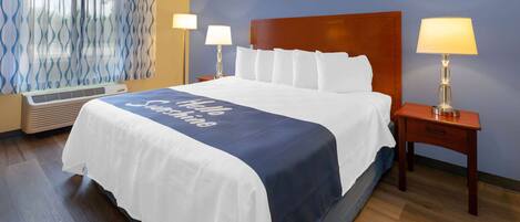 A hotel room with a large bed, two bedside tables with lamps, a window with curtains, and a wall air conditioner.