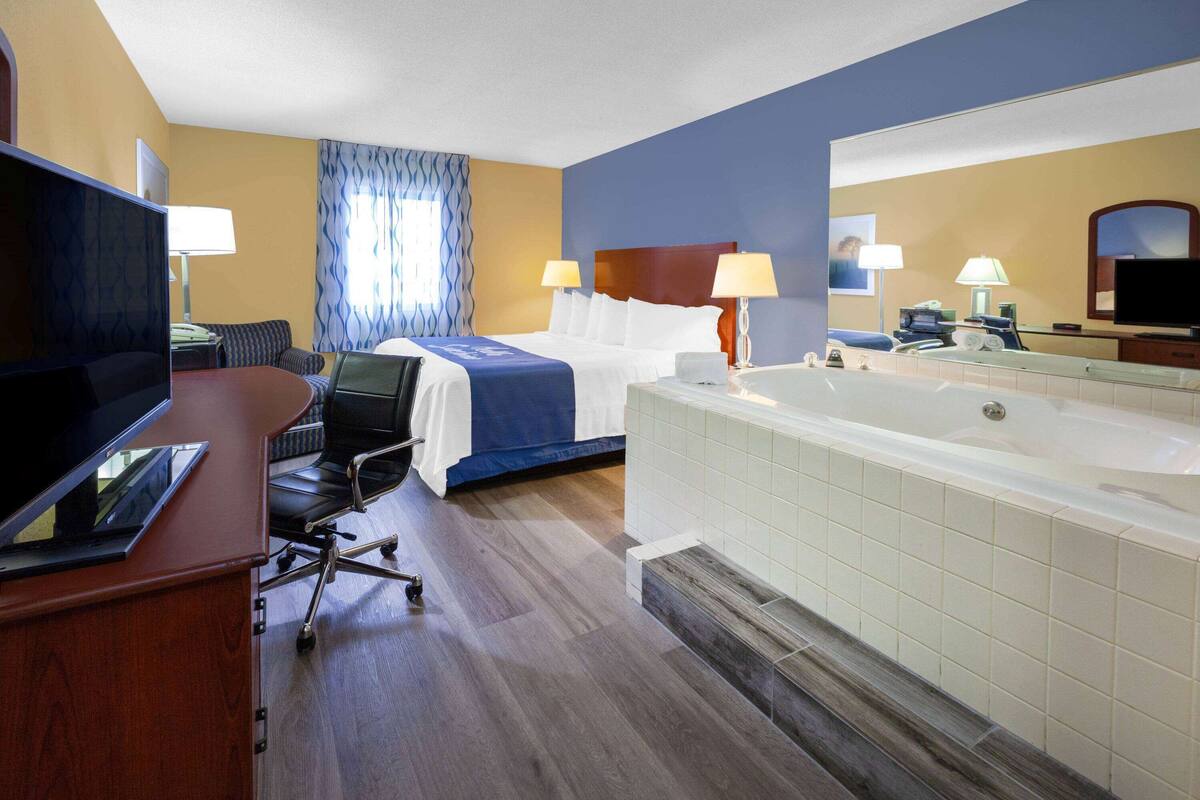 A hotel room with a bed, a desk with a computer, a chair, a large bathtub, and a television.