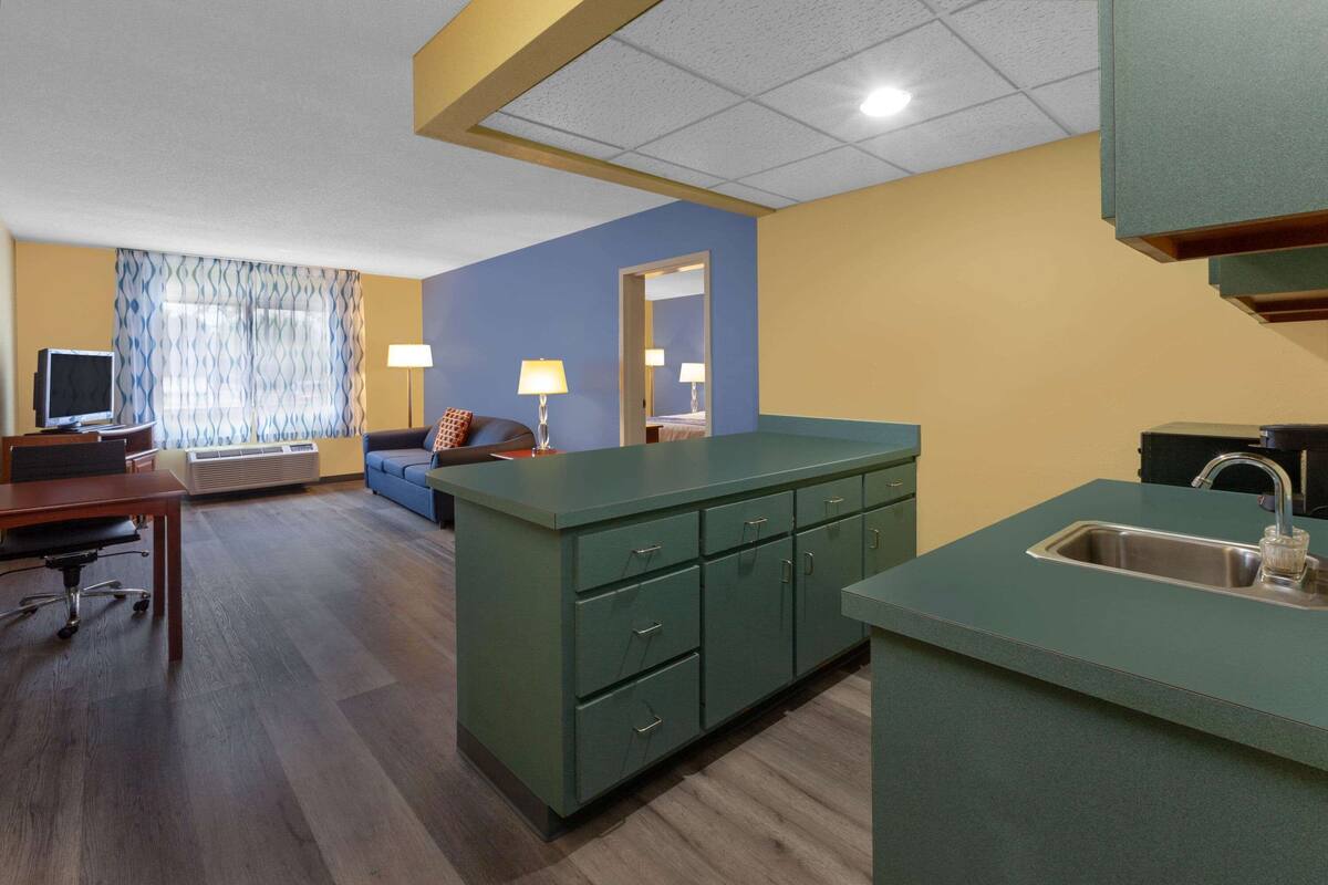 A compact hotel room with a kitchenette, a sofa, a desk, and a TV.