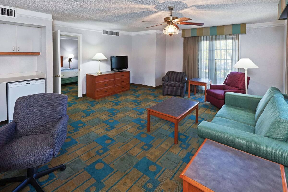 A hotel room with a desk, a television, a sofa, a chair, and a ceiling fan.